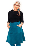 A woman with long gray hair in a braid, wearing glasses and a black top, stands smiling against a white background, modeling the BUTIK KUBIK Cissi och Selma Sonja skirt in petrol with pockets.