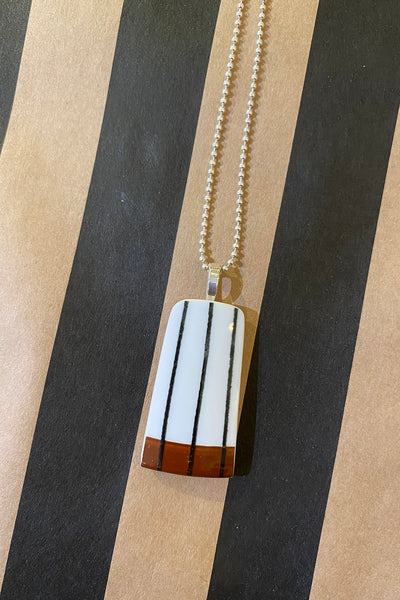 Saved & remade necklace Porslin by BUTIK KUBIK: A rectangular pendant crafted from återbrukat porslin, with black stripes and a brown band, hangs on a silverpläterad kedja against black and tan striped background.