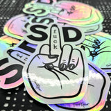 A stack of Favoretro sticker Fuck SD by BUTIK KUBIK: holographic vinyl stickers featuring a hand with painted nails flipping the bird. “Fuck” runs along the finger, bold S and D on either side—an eye-catching political statement.