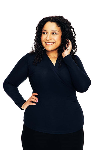 A woman with curly hair smiles while looking to the side, wearing a Cissi och Selma blus Susanne Marinblå from BUTIK KUBIK, posing with one hand on her hip and the other touching her hair against a white background.