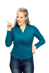 Smiling woman with long gray hair wearing the Cissi och Selma blus Susanne Petrol from BUTIK KUBIK and jeans points upward with one finger against a white background.