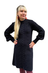 A woman with long blonde hair wears the Svea Kol dress by BUTIK KUBIK, featuring balloon sleeves, along with red earrings and mauve tights. She stands with hands on hips and smiles against a white background.