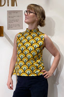 A person confidently models the Kubik top cineast Bridges by BUTIK KUBIK, featuring a 60s inspired sleeveless design with vibrant green, yellow, orange, and brown scallops. Made from GOTS certified organic cotton, they have short hair and glasses against a white wall with partially visible text.