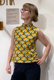 A person confidently models the Kubik top cineast Bridges by BUTIK KUBIK, featuring a 60s inspired sleeveless design with vibrant green, yellow, orange, and brown scallops. Made from GOTS certified organic cotton, they have short hair and glasses against a white wall with partially visible text.