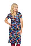 A woman with long blonde hair poses in the Cissi och Selma klänning Betty Rödvinge by BUTIK KUBIK, a blue V-neck dress with a vibrant floral and butterfly print, standing against a white background.