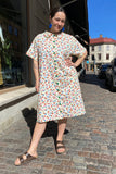 A woman smiles on a sunny cobblestone street, wearing the Kubik blouse dress vintage cotton by BUTIK KUBIK, styled with brown sandals and blue earrings. She poses confidently with one hand on her hip and her head slightly tilted.