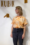 A blonde woman with glasses stands by a white wall displaying "KUBIK" and wooden decor, wearing the BUTIK KUBIK Kubik blouse vintage—an upcycled peach-orange patterned piece—with dark jeans, looking to her left.