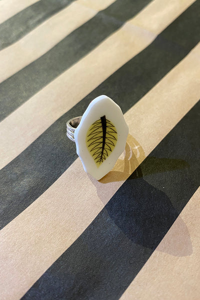The Saved & Remade ring porslin by BUTIK KUBIK is a silverplated ring with a white, hexagonal face of recycled porcelain featuring a black leaf design, shown on a beige and black striped surface with a distinct shadow.