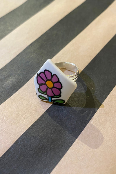 The Saved & remade ring porslin by BUTIK KUBIK is a silverplated ring with a pentagon-shaped face of recycled porcelain, featuring a colorful cartoon flower on striped black and beige.