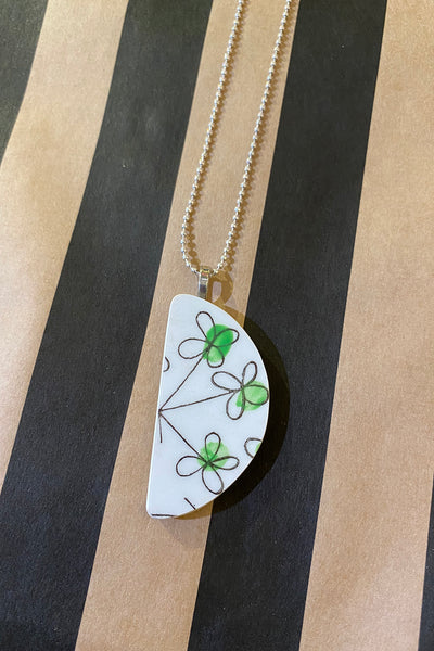 The Saved & remade necklace Porslin from BUTIK KUBIK features a half-moon pendant of återbrukat porslin with green and black clovers on wide black and brown stripes, finished with a silverpläterad kedja.