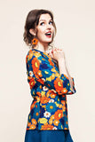 A woman wearing the Cissi och Selma top Vendela 1970 Blåorange from BUTIK KUBIK, featuring a colorful floral pattern, and orange flower earrings, smiles with hands clasped and looks upward against a plain light background.