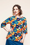 A woman wearing the Cissi och Selma top Vendela 1970 Blåorange from BUTIK KUBIK, featuring a colorful floral design in orange, yellow, and blue, poses with one hand on her hip and smiles at the camera. She accessorizes with orange earrings and has wavy brown hair.