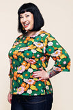 Smiling woman with a dark bob haircut wearing the BUTIK KUBIK Cissi och Selma top Vendela 1970 Grönorange—a colorful green, orange, and pink floral A-line top with three-quarter sleeves—paired with dark jeans and red nail polish, standing sideways with one hand on her hip against a plain background.