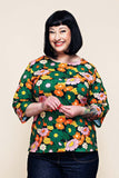 A woman with a dark bob haircut smiles, wearing the Cissi och Selma top Vendela 1970 Grönorange from BUTIK KUBIK—a green A-line top with a colorful floral pattern and three-quarter sleeves—paired with dark jeans, standing against a plain background.