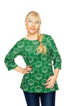 A woman with blonde hair in a side braid, wearing the Cissi och Selma top Vendela Retroblom Grön from BUTIK KUBIK—a green stretch cotton jersey top with a white floral pattern—smiles with one hand on her hip against a white background.