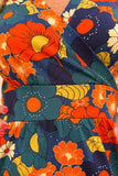 A close-up of the Cissi och Selma dress Vera 1970 Blåorange by BUTIK KUBIK, a vibrant bomullstrikå dress featuring a floral pattern in orange, blue, yellow, and white on navy blue fabric. This klänning Vera showcases a wrap-style bodice and gathered waist details.
