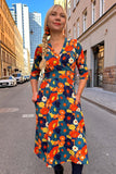 A woman with a long blonde braid wears the Cissi och Selma dress Vera 1970 Blåorange from BUTIK KUBIK—a colorful, floral-patterned omlottklänning with pockets—while standing on a city street lined with tall buildings and parked cars.