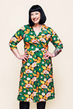 A smiling woman with short black hair wears the Cissi och Selma dress Vera 1970 Grönorange by BUTIK KUBIK, featuring a colorful floral pattern and practical pockets; she stands with her hands on her hips against a light background.
