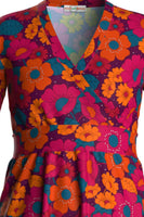 Introducing the Cissi och Selma dress Vera Humlesurr Magenta from BUTIK KUBIK, a vibrant wrap dress featuring a V-neckline. This organic cotton piece displays a bold floral pattern in orange, pink, teal, and yellow with long sleeves and a gathered waist for added femininity.