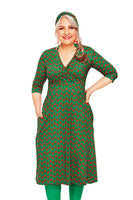 A woman with long hair stands confidently in a Cissi och Selma Vera Sekund Grön dress by BUTIK KUBIK, featuring a green V-cut design with red and orange circles. She pairs it with green tights and a matching headband, hand on her hip, smiling against a white background.