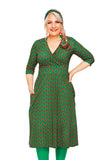 A woman with long hair stands confidently in a Cissi och Selma Vera Sekund Grön dress by BUTIK KUBIK, featuring a green V-cut design with red and orange circles. She pairs it with green tights and a matching headband, hand on her hip, smiling against a white background.