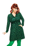 A red-haired woman in a ponytail wears the BUTIK KUBIK Cissi och Selma dress Viveca Paraply grön with green tights, earrings, and bright red lipstick, standing against a white background.