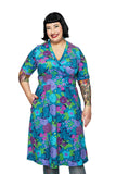 Smiling woman with short black hair and visible arm tattoos wears the BUTIK KUBIK Cissi och Selma Vivi dress Trädgård lila—a bright blue dress with a colorful floral pattern and pockets—standing with one hand in her pocket against a white background.