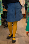 Indoors, a person wears the BUTIK KUBIK Kubik corduroy zip skirt nr 2 in blue, paired with yellow tights and black Mary Jane shoes, with colorful Kubik clothing and wooden flooring seen in the background.