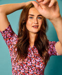 A person with long brown hair and blue eyes poses against a blue background, adorned in a floral Jumperfabriken Dress Maria Pink by BUTIK KUBIK. The wrap design enhances their charm as they gracefully raise an arm, partially covering their face while smiling gently.