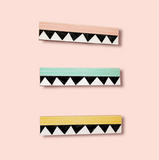 Three Törmi Iiris hair clips from BUTIK KUBIK, made of Finnish Birch plywood, are vertically arranged against a pink background. The clips are pink, mint green, and yellow with a black and white triangular pattern.