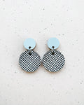 Two Törmi hento earrings by BUTIK KUBIK with small, solid light blue circles on top and larger, dangling circles below featuring a black and white crisscross pattern, crafted from Finnish birch plywood and displayed on a speckled background.