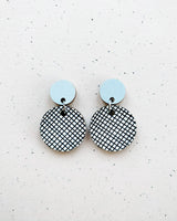 Two Törmi hento earrings by BUTIK KUBIK with small, solid light blue circles on top and larger, dangling circles below featuring a black and white crisscross pattern, crafted from Finnish birch plywood and displayed on a speckled background.