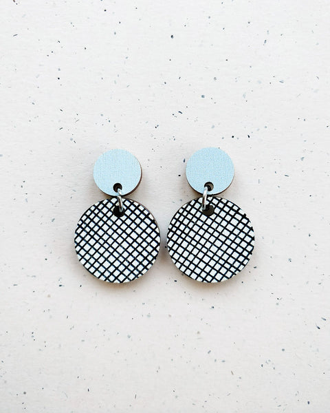 Two Törmi hento earrings by BUTIK KUBIK with small, solid light blue circles on top and larger, dangling circles below featuring a black and white crisscross pattern, crafted from Finnish birch plywood and displayed on a speckled background.