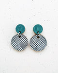 A pair of BUTIK KUBIK Törmi hento earrings petrol, with small green studs and large white pendants with a black crisscross pattern, are displayed on a speckled white background.
